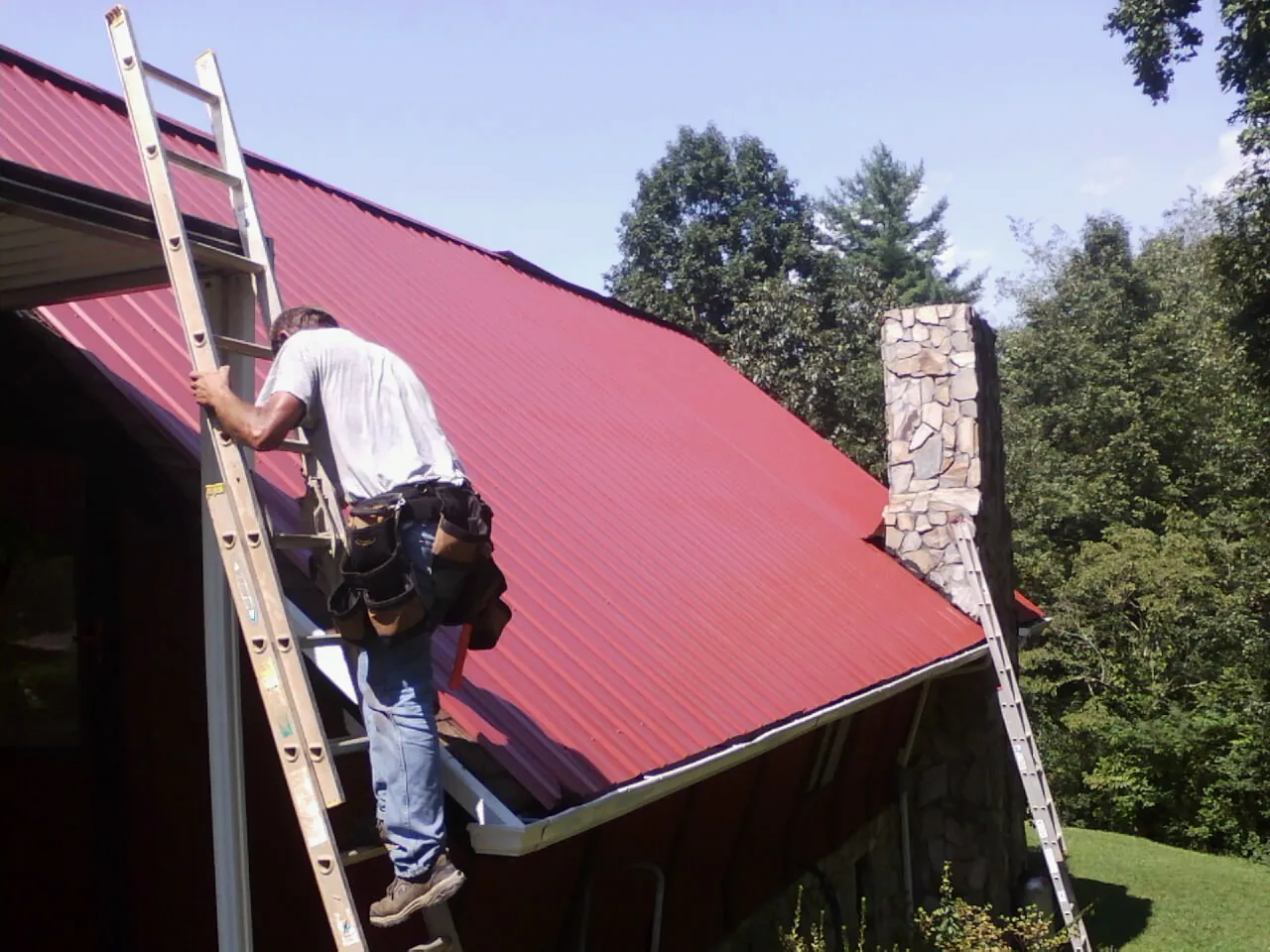 Roofer on a ladder beside a red metal roof and stone chimney — Soffit & Fascia Repair by Roofing Brothers in Limekiln